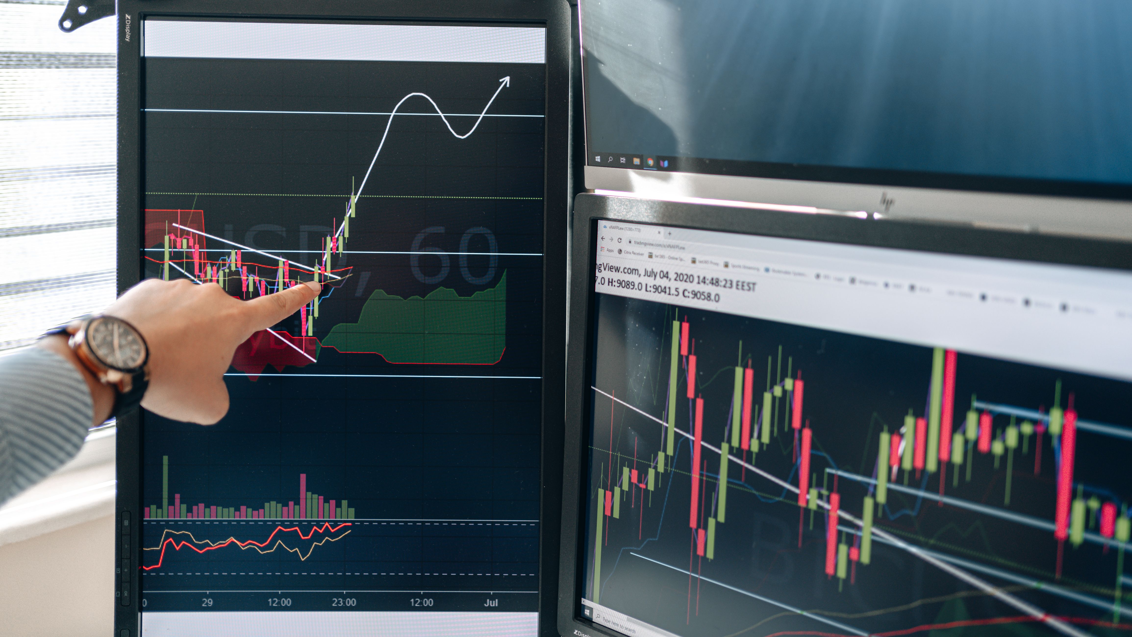 Project Categories Person pointing at stock market graphs on dual monitors in a modern workspace.